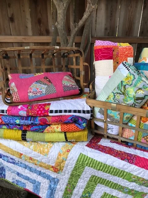 Several folded quilts in bright pinks, yellows, greens and blues, stacked in wooden baskets outdoors in front of a plank fence