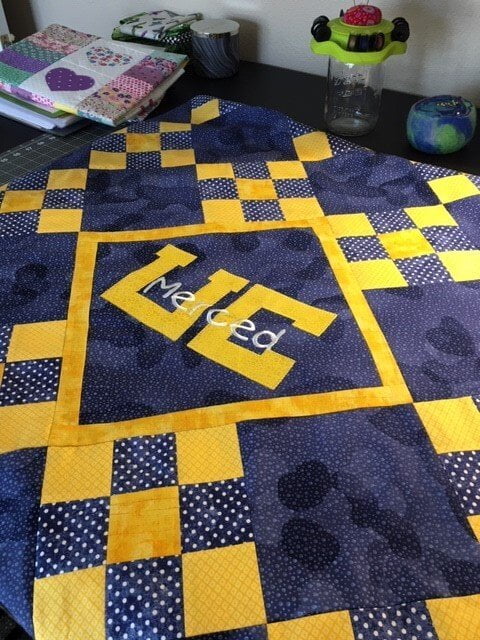 A navy and yellow quilt with a large yellow UC and the word Merced in white script, folded on a table beside other quilts