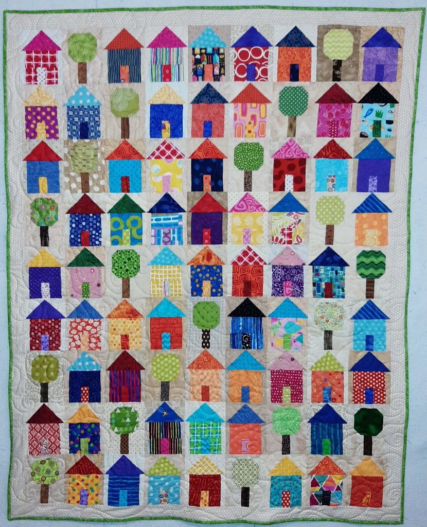 A quilt of small patchwork houses and trees on a cream background, hung flat against a white wall