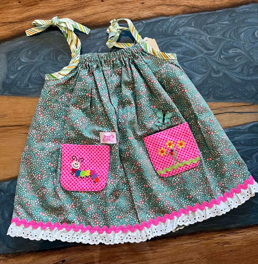 A finished child's dress laid flat: green and yellow striped tie straps, sage green floral fabric, two hot pink polka-dot pockets with embroidered flowers, a caterpillar and a butterfly, and pink trim at the hem