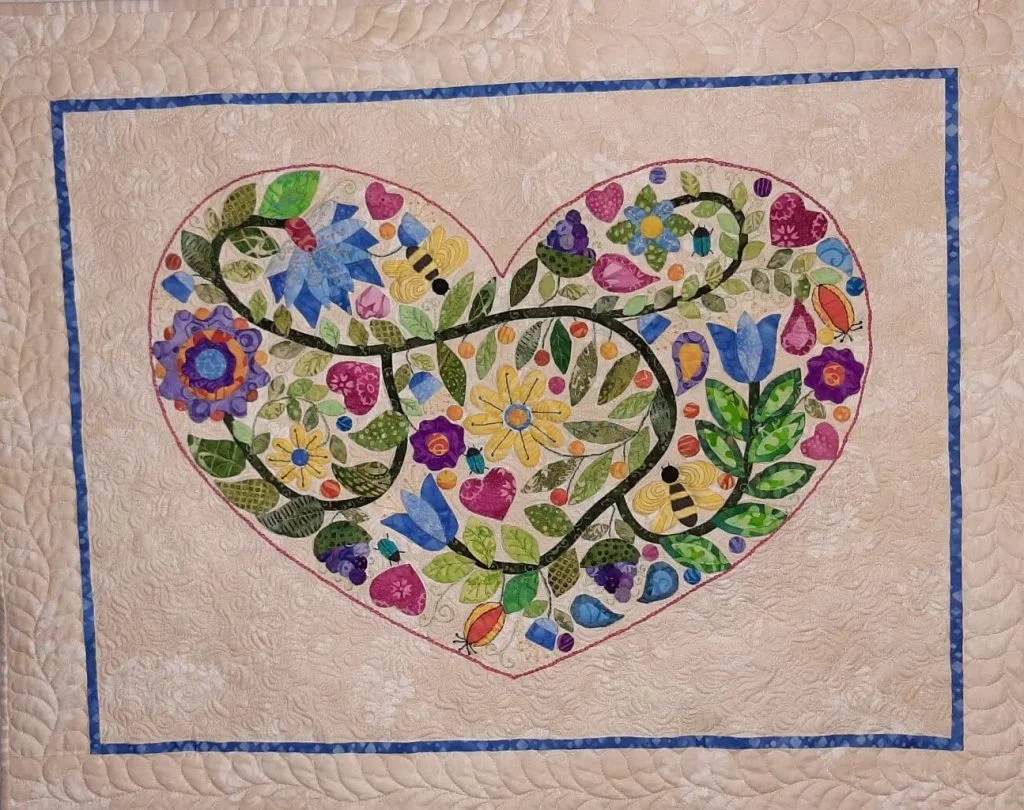 A cream quilt with an appliqued heart of green vines, leaves and flowers, edged with a thin pink line inside a royal blue border